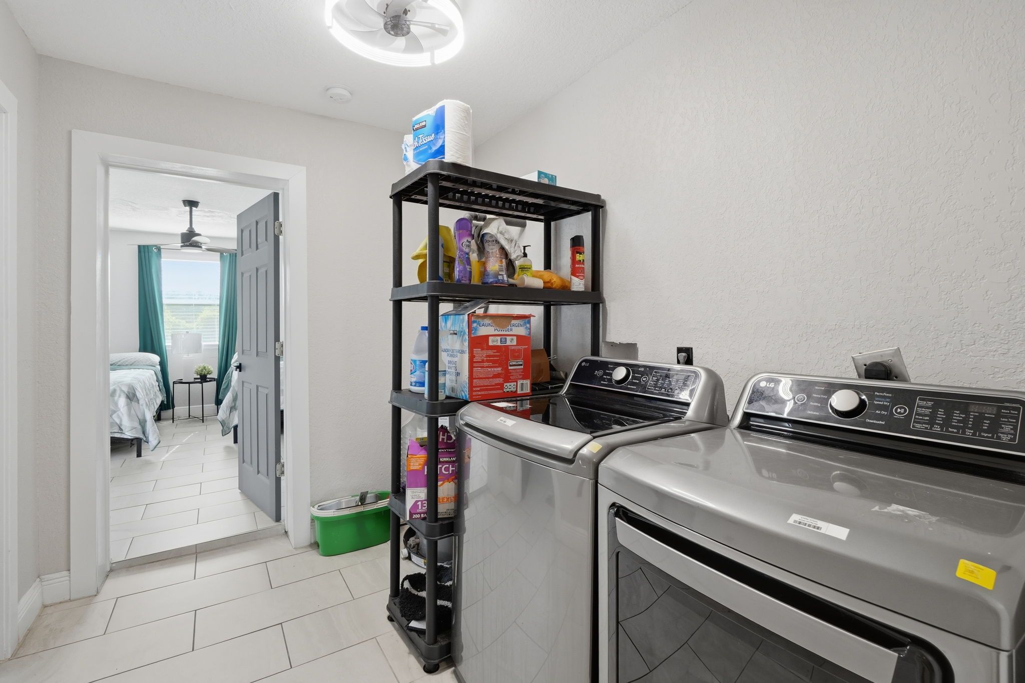 Un espace buanderie comprenant un lave-linge et un sèche-linge, une étagère avec des produits de nettoyage et une porte donnant sur une chambre.