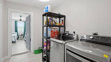 Un espace buanderie comprenant un lave-linge et un sèche-linge, une étagère avec des produits de nettoyage et une porte donnant sur une chambre.
