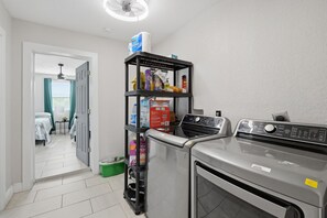 A laundry area with a washing machine and dryer, a shelf with cleaning supplies, and a door leading to a bedroom.