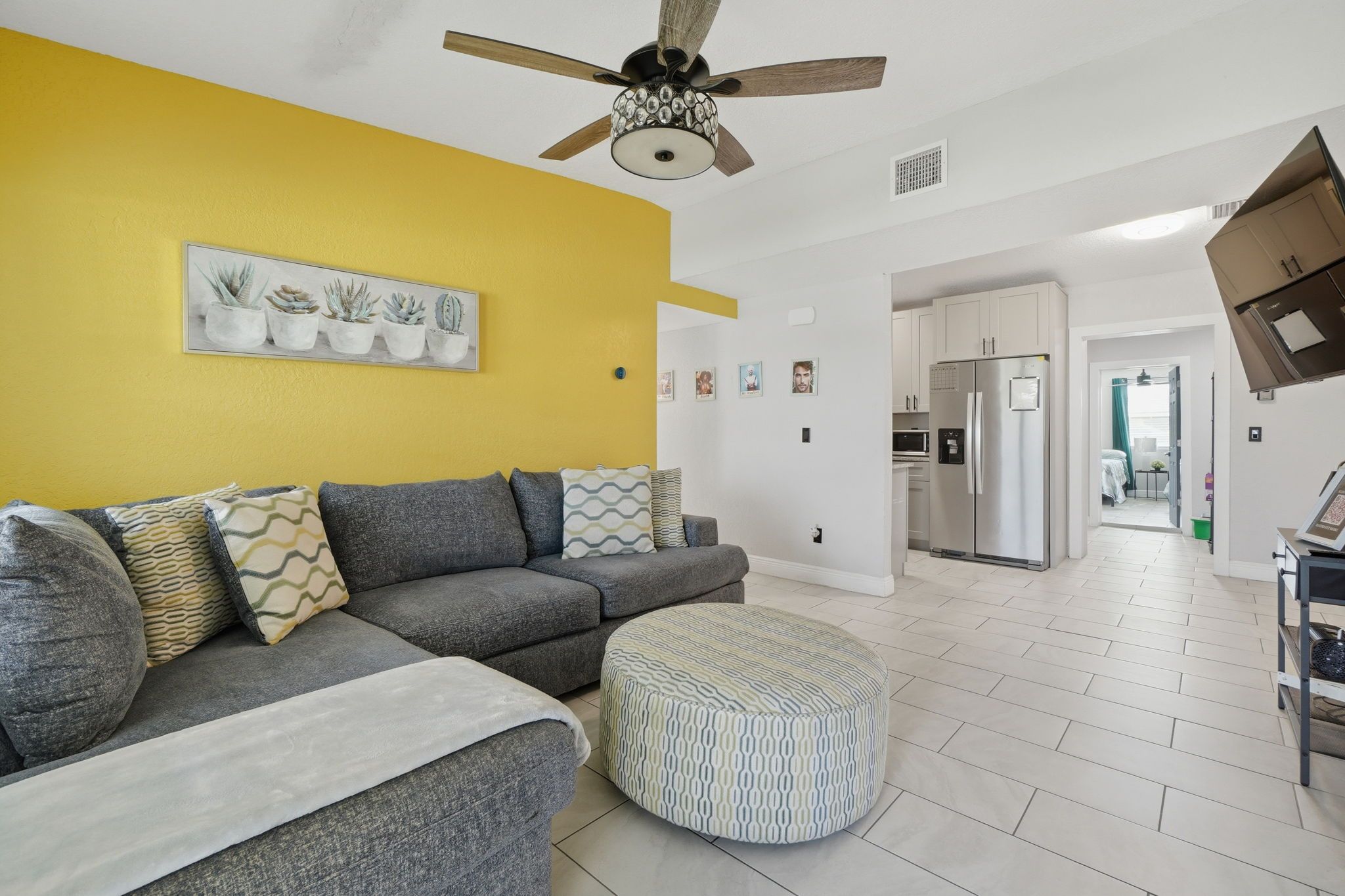 A living room with a sofa, ottoman, ceiling fan, and a kitchen in the background.