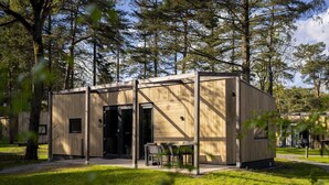 A modern, single-story wooden cabin with a glass door entrance, a small patio with a table and chairs, surrounded by a grassy area and trees. - Cozy Glamping in Mierlo (Mierlo)