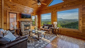 A cozy cabin interior with a stone fireplace, leather sofa, and large windows offering mountain views.
