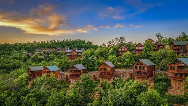 A cluster of log cabins with multiple stories, surrounded by dense forest, and set against a backdrop of a sunset sky.