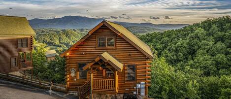 A log cabin with a porch, a sign indicating it is a historic site, and a view of a forested mountain range in the background.
