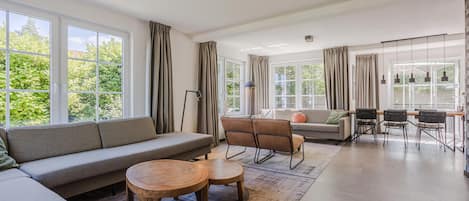 En rummelig opholdsstue med sofa, stole, et sofabord og et spiseområde med stole og bord. Store vinduer giver naturligt lys og udsigt til grøn bevoksning udenfor.