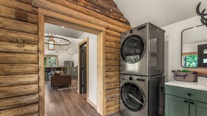 Un área de lavandería con lavadora y secadora apiladas, paredes de madera y vista a una sala de estar con sofá y candelabro.