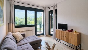A modern living room with a grey sofa, a wooden TV stand, and a large window offering a view of greenery. - Serenity Apartment (Posada De Llanes)