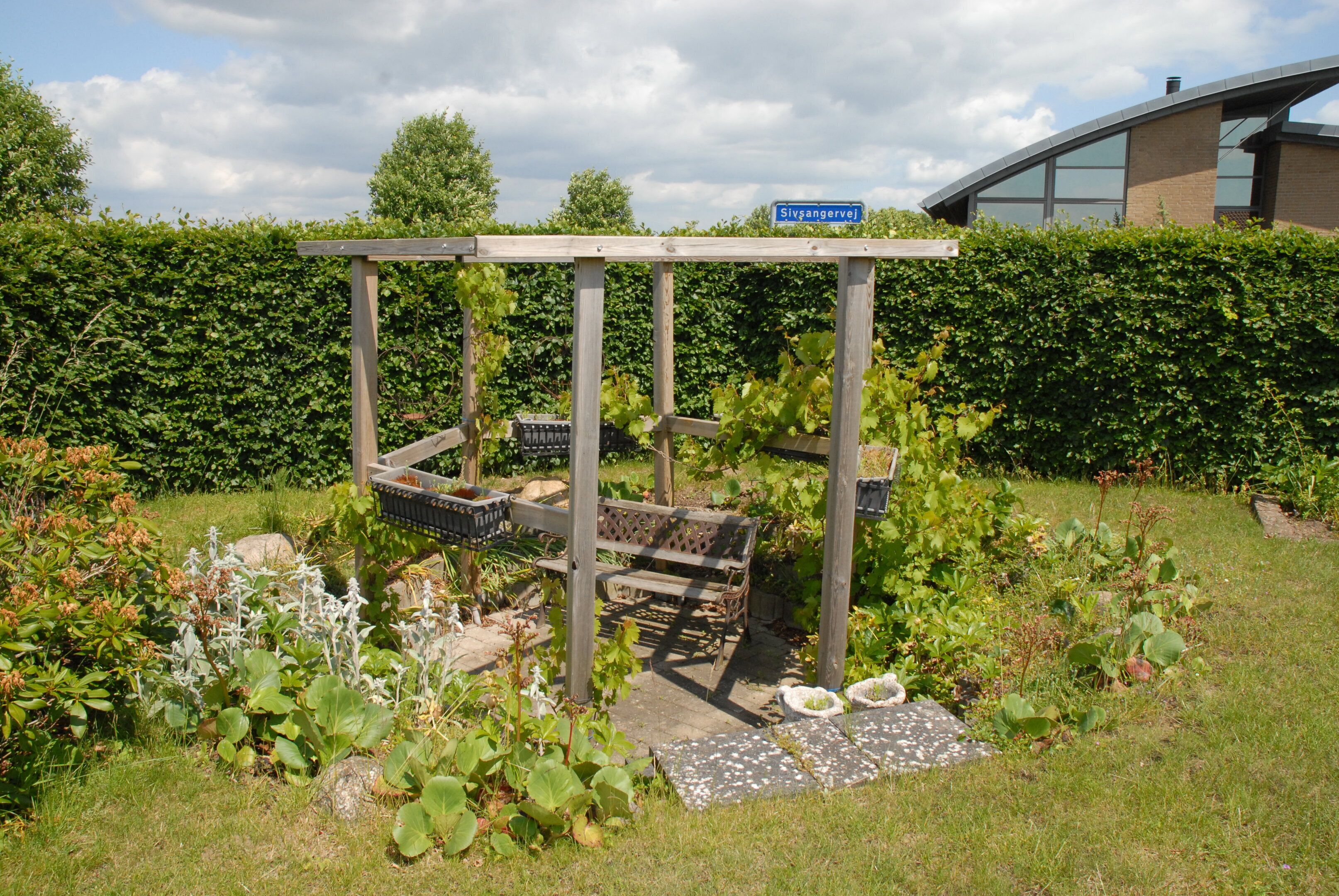 Ein Garten mit einer Holz-Pergola, einer Bank und einem Schild mit der Aufschrift 'Stivangervej'.