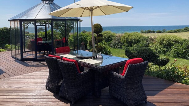 En pavillon med glasvægge, et bord, stole og en parasol, med udsigt over havet.