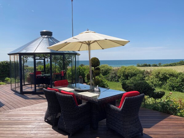 A glass-enclosed gazebo with a table and chairs, a large umbrella, and a view of the sea.