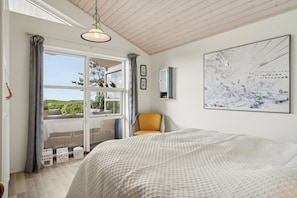 A bedroom with a large bed, a window with curtains, a chair, and a wall-mounted mirror. - Fantastic summer house with sea view (Ebeltoft)