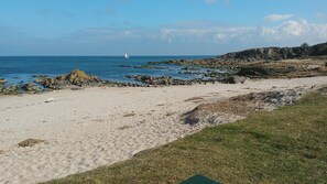 Una playa arenosa con afloramientos rocosos, un velero a lo lejos y una zona cubierta de césped al frente.