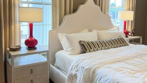 Een net opgemaakt bed met witte lakens en een decoratief kussen, geflankeerd door twee nachtkastjes met lampen, in een kamer met grote ramen en gordijnen.