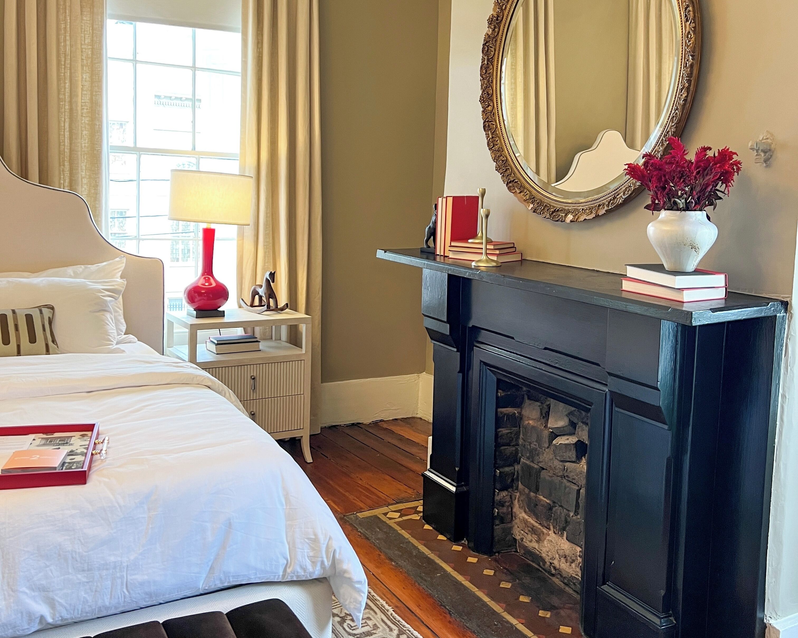 A hotel room with a bed, a fireplace, a mirror, and a nightstand with books and a lamp.