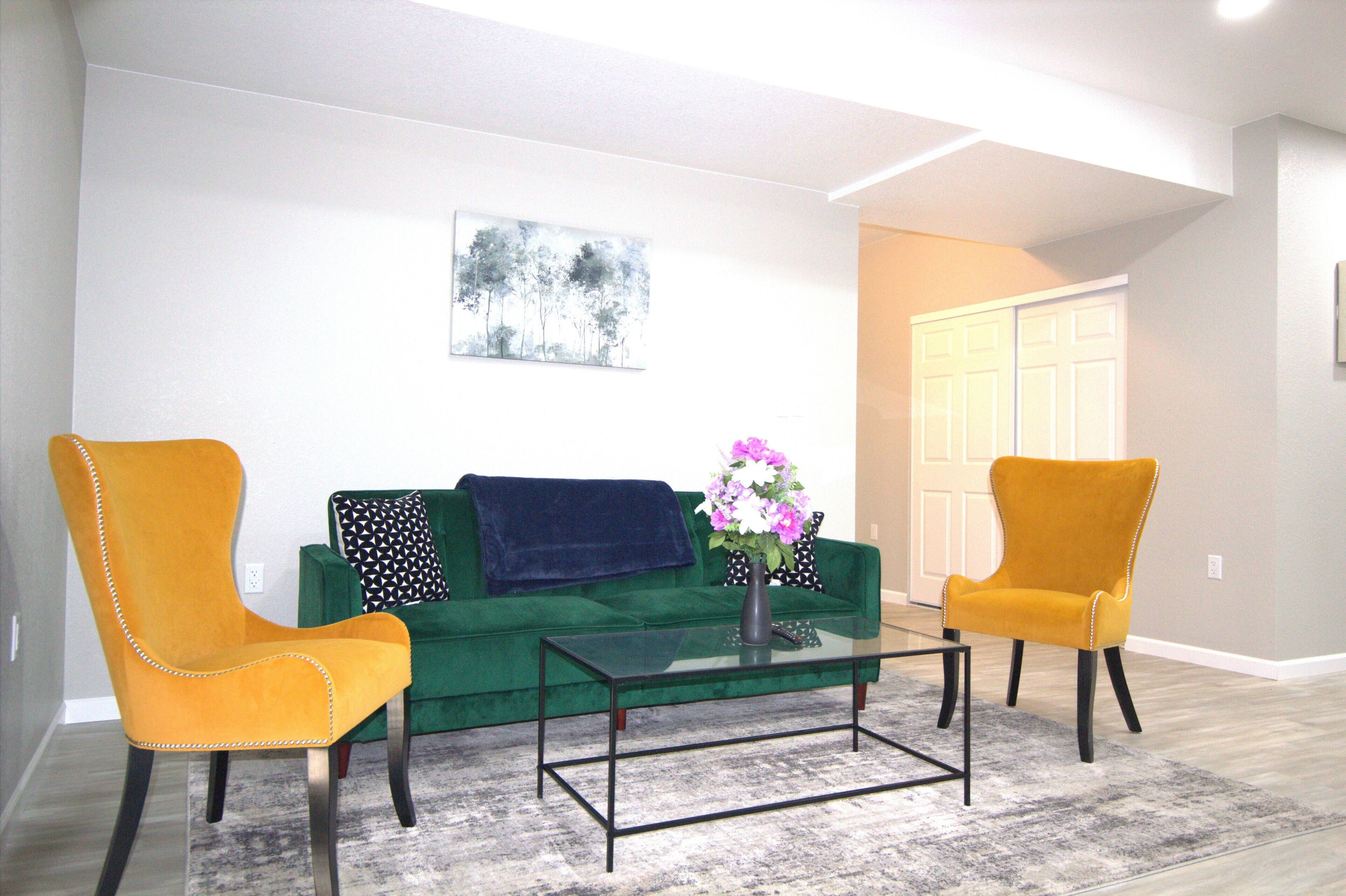 A living room with a green sofa, two yellow chairs, a glass coffee table, and a vase with flowers.