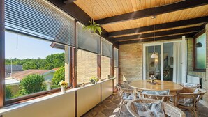 Eine überdachte Terrasse mit rundem Tisch und Korbstühlen, große Fenster mit Jalousien und Blick ins Viertel.
