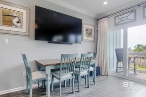 Un comedor con mesa de madera y sillas azul claro, televisor de pantalla plana en la pared y un balcón con vistas.