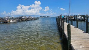 Un port de plaisance avec des bateaux amarrés à des quais en bois, un ciel bleu clair et des bâtiments qui se dessinent à l’horizon.