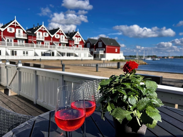 A coastal view with a row of red and white buildings, two glasses of red wine, and a potted plant on a deck.