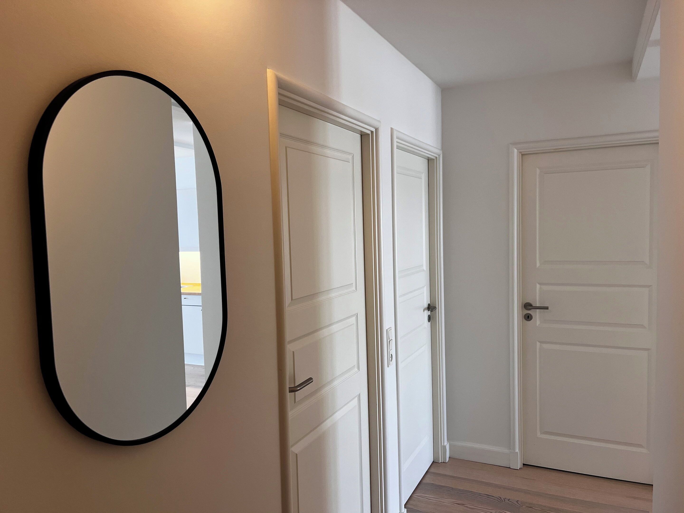 Un pasillo con puertas blancas, un espejo redondo y suelo de tonos claros.