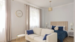 A bedroom with a bed, a sofa, a bedside table, a lamp, a clock, and a window with curtains. - Chmielna Green Apartment (Warszawa)