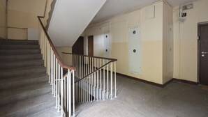 A staircase with a wooden handrail and white balusters, leading to a landing with beige walls and a door with a vent. - Chmielna Green Apartment (Warszawa)