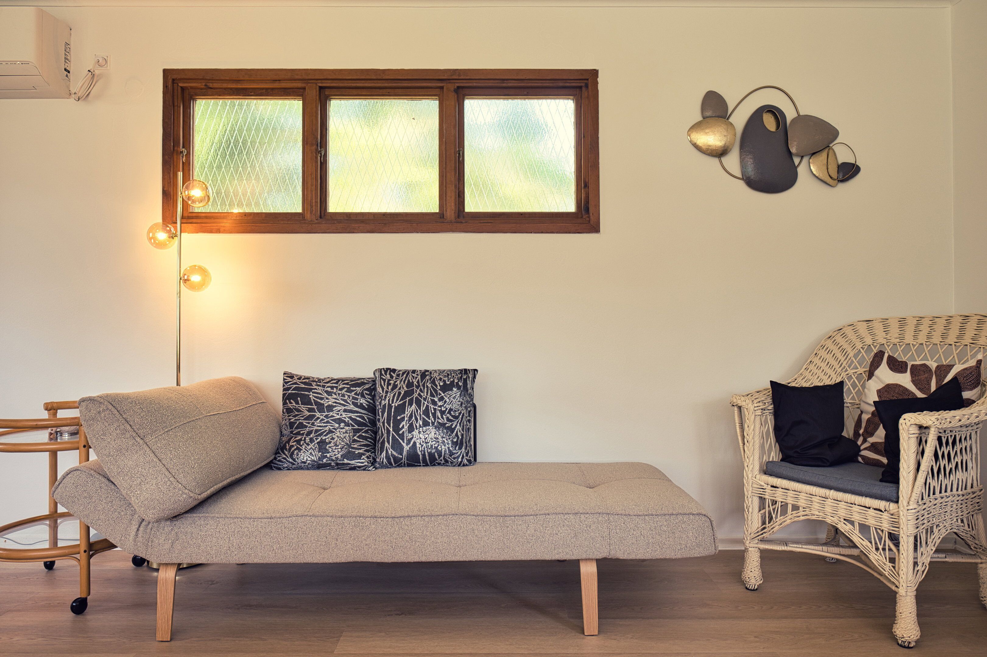 Een kamer met een bank, een rieten stoel, een raam met jaloezieën en een wandlamp.