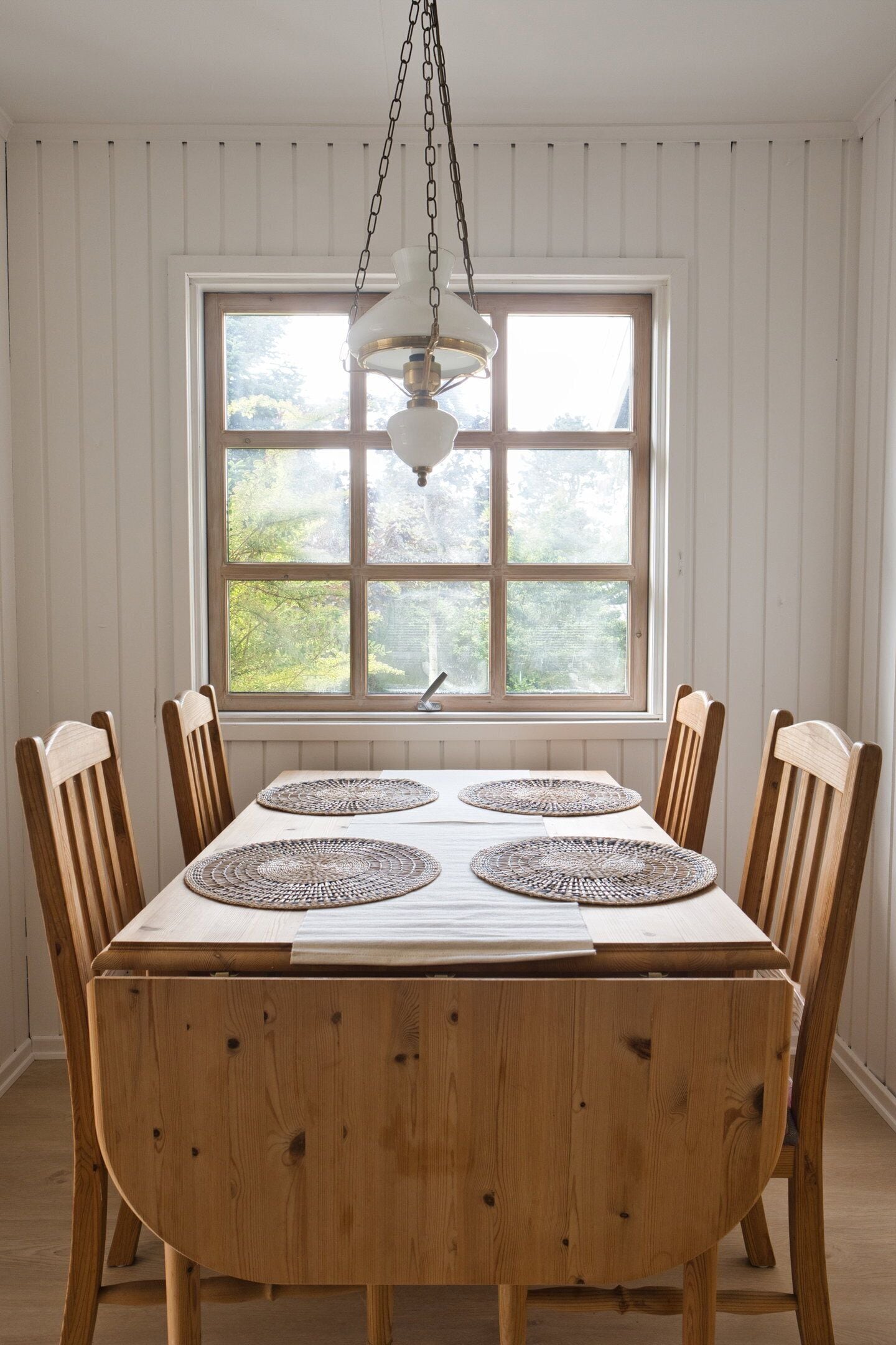 A dining table with four placemats, wooden chairs, a hanging light fixture, and a window with a view of greenery.