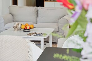 Un salon avec un canapé, une table basse sur laquelle se trouve un saladier rempli de fruits et des bougies, ainsi qu’un magazine.
