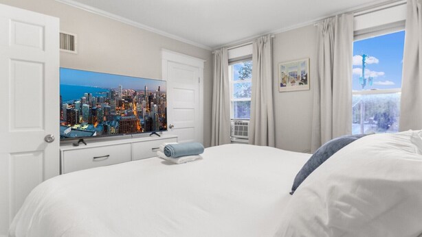 Una habitación de hotel con cama, televisor mostrando un paisaje urbano, ventana con cortinas y un pequeño equipo de aire acondicionado.