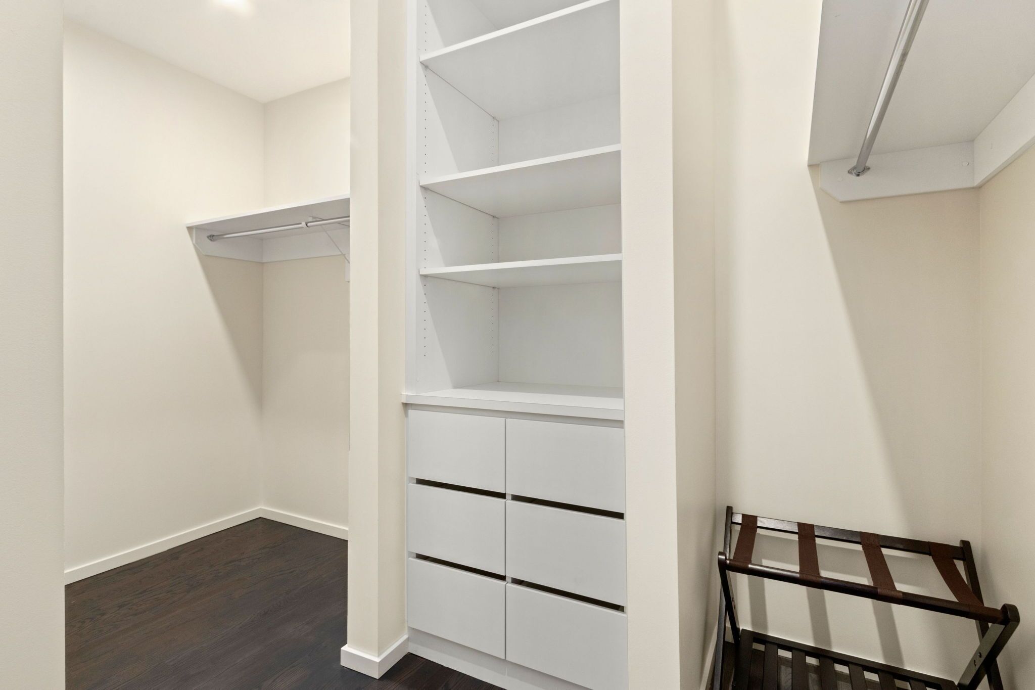 A walk-in closet with white shelving and drawers, a metal rack, and a dark wooden floor.