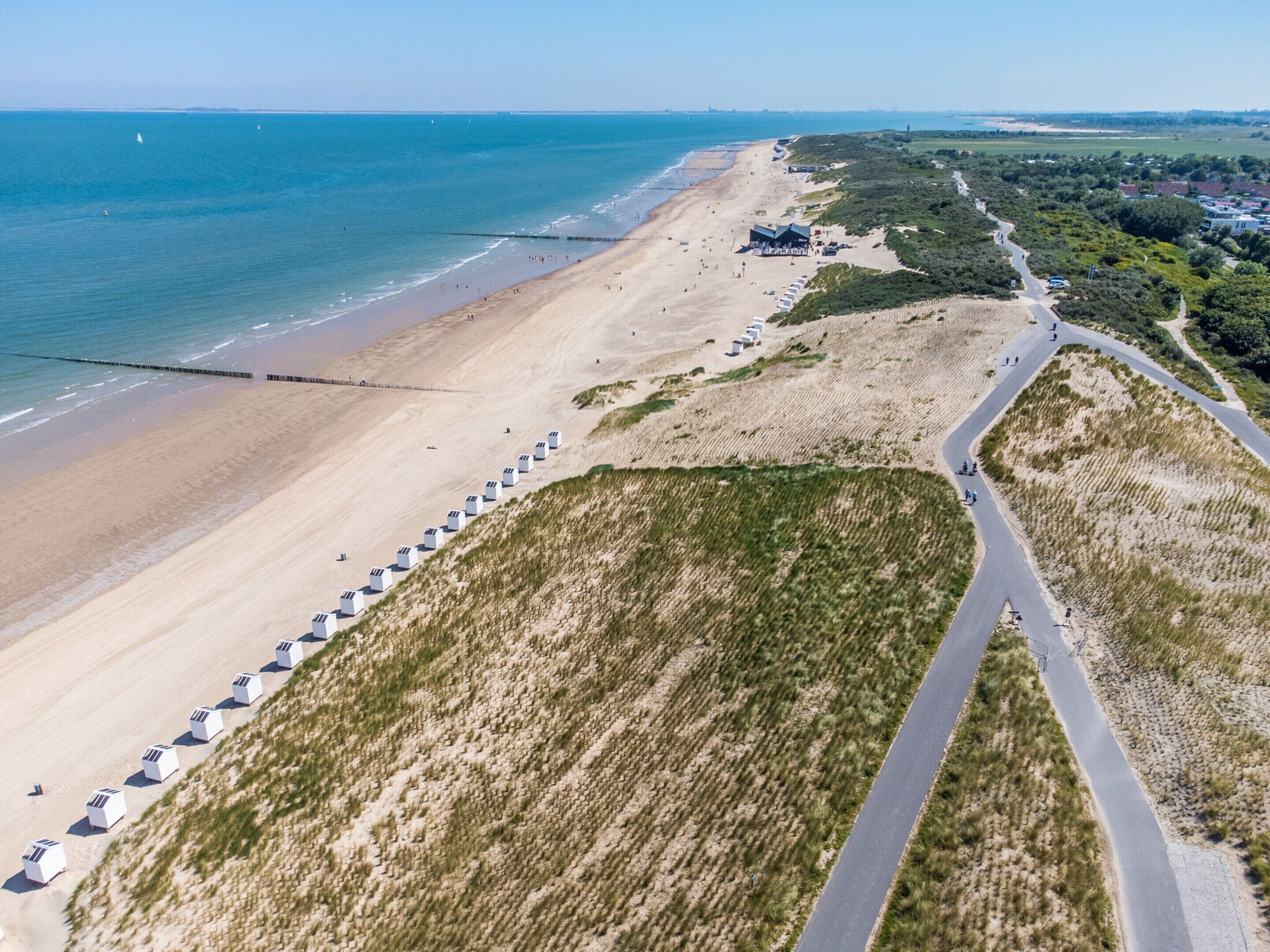 An aerial view of a beach with a row of beach huts, a sandy beach, and a road curving through the dunes.