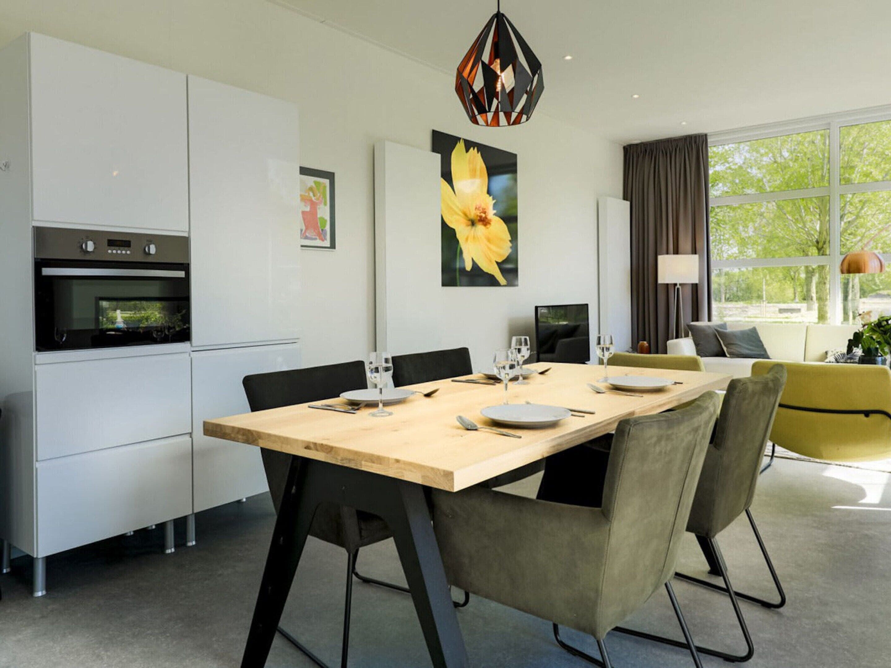 A modern kitchen with built-in appliances, a dining area with a wooden table and chairs, and a large window with a view of greenery.