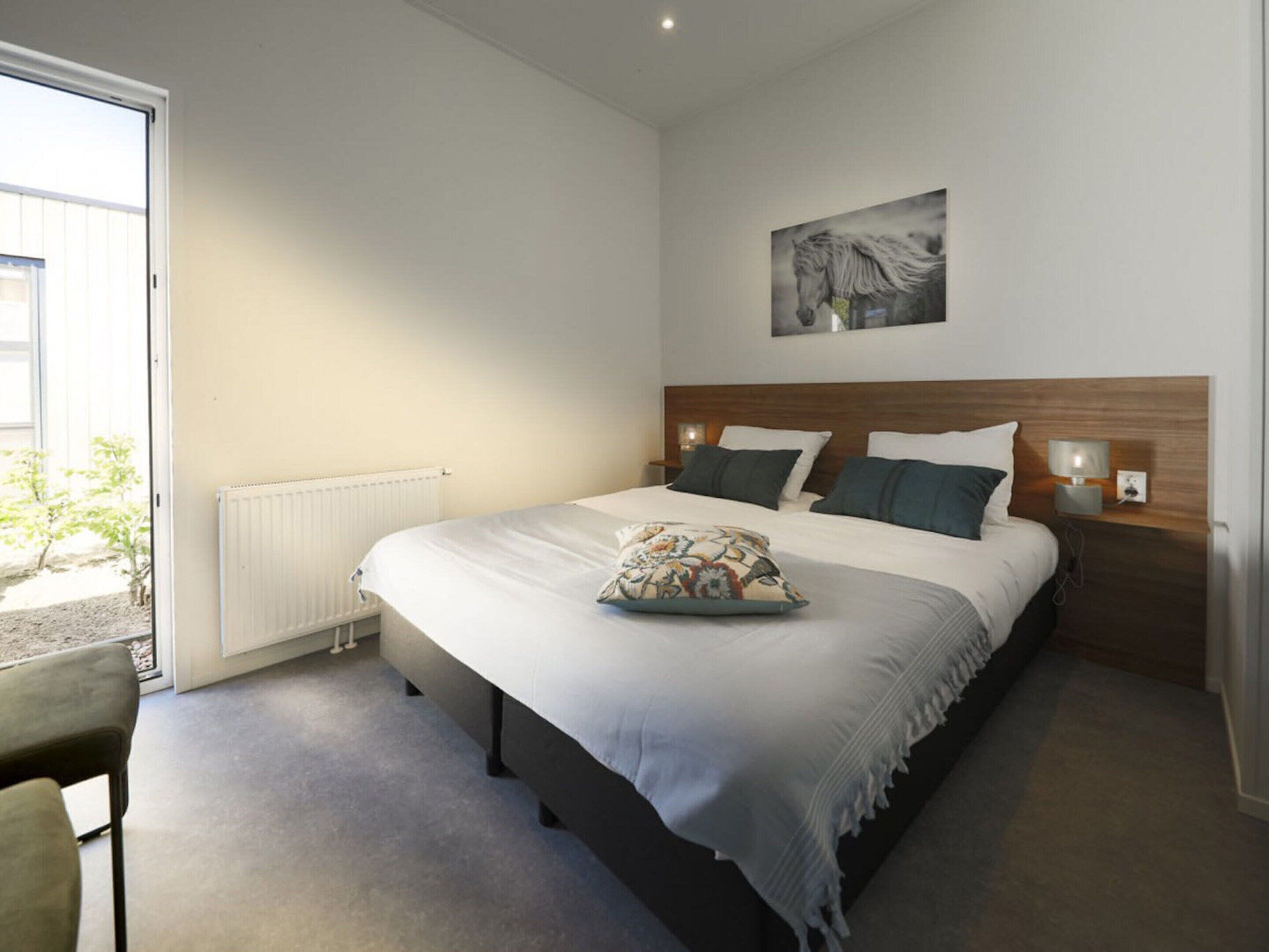 A bedroom with a large bed, a headboard, bedside lamps, a radiator, a chair, and a window with a view of greenery.