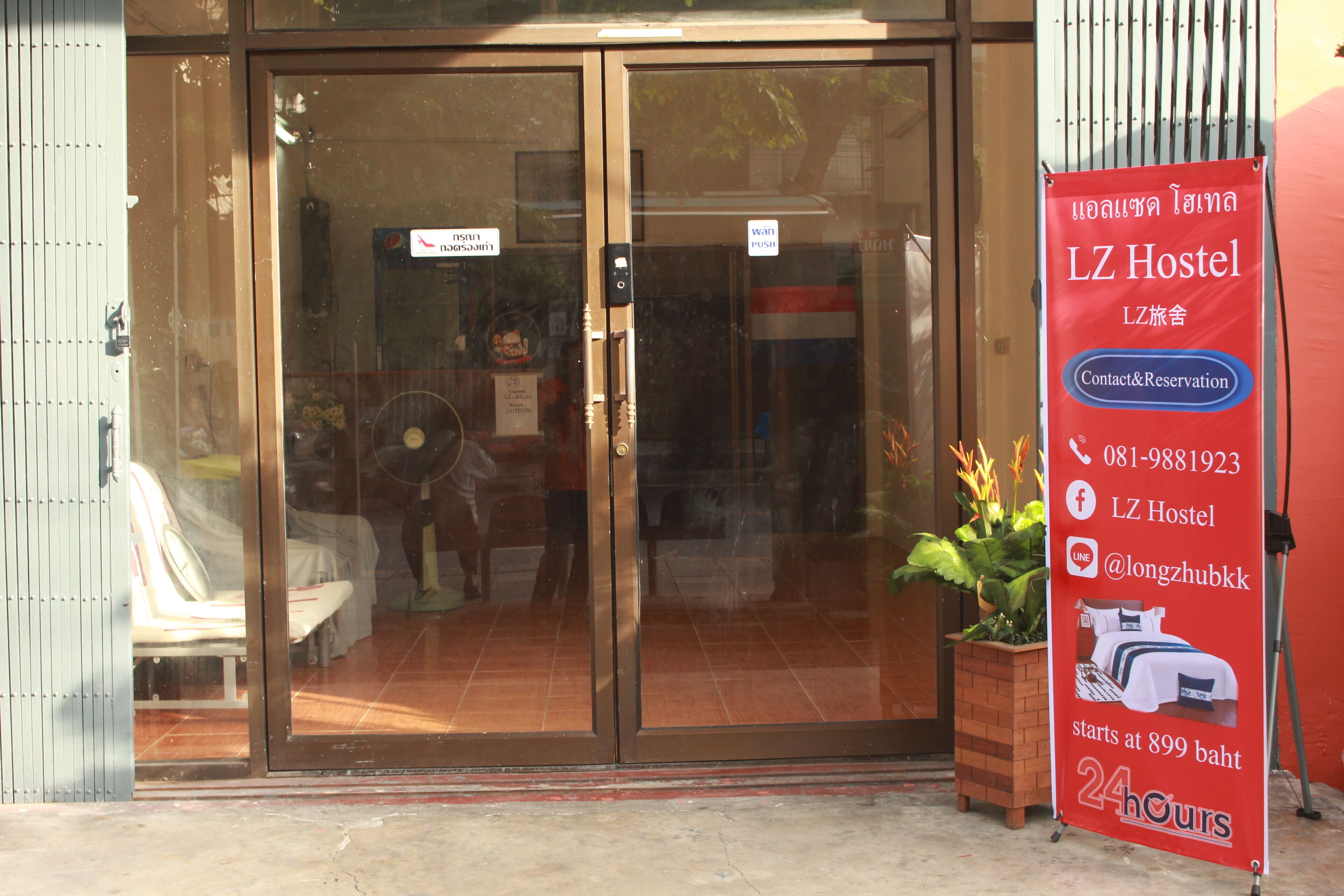 A hostel entrance with a glass door, a signboard advertising the hostel, and a potted plant outside.