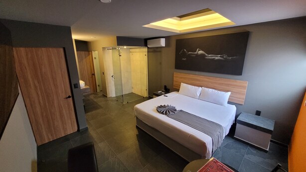 A modern hotel room with a large bed, a bedside table, a nightstand, a wall-mounted TV, and a shower area. - 4Play Motel (El Marques)