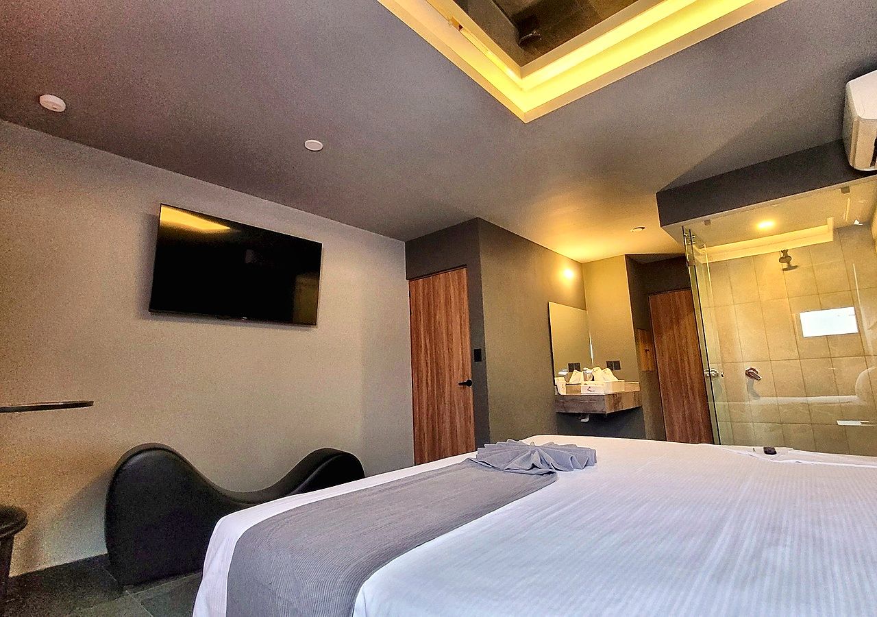 A hotel room with a bed, a desk, a chair, a TV, a mirror, and a shower area.