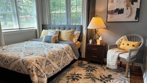 A bedroom with a large bed, a nightstand, a chair, and a window with blinds. - Charming 3-Bedroom Yellow Ranch Retreat near Lake Michigan (Benton Harbor)