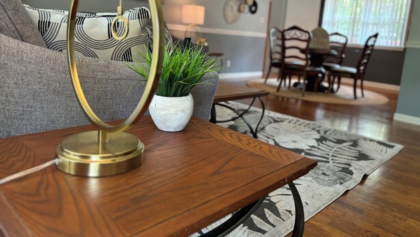 A living room with a wooden coffee table, a white pot with green plants, a grey sofa with patterned cushions, and a dining area in the background. - Charming 3-Bedroom Yellow Ranch Retreat near Lake Michigan (Benton Harbor)