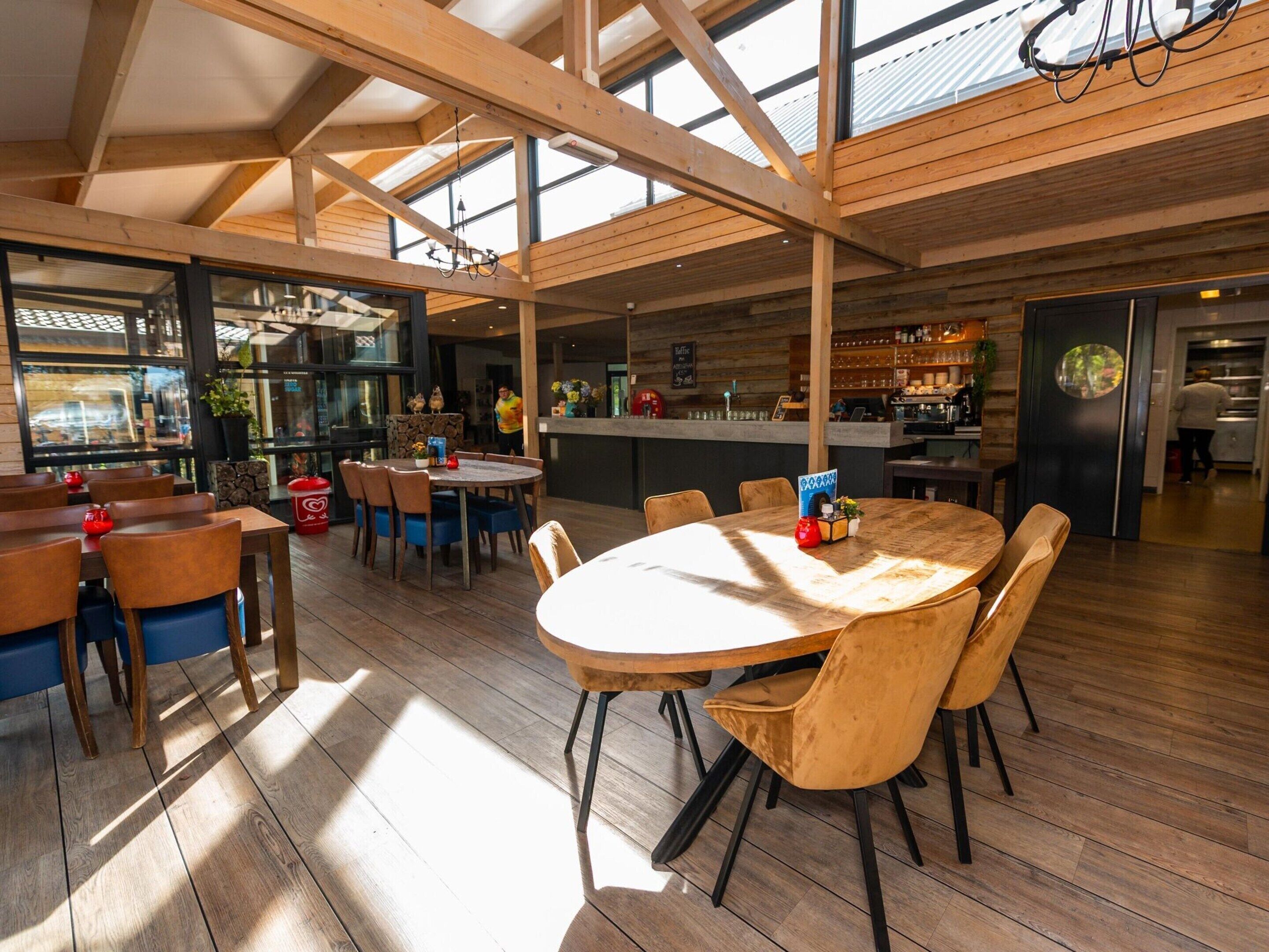 A spacious dining area with wooden tables and chairs, a kitchen in the background, and large windows allowing natural light.