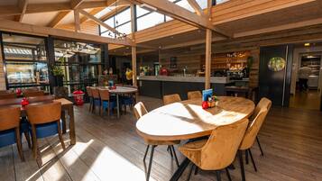 A spacious dining area with wooden tables and chairs, a kitchen in the background, and large windows allowing natural light.