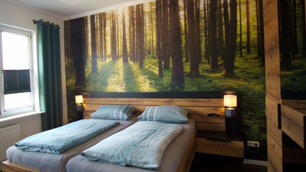 A double bed with a forest mural on the wall, a window with curtains, and a bedside table with a lamp. - Holiday Apartment Busch (Bad Sachsa)