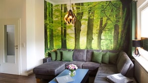 A modern living room with a sectional sofa, a blue coffee table, and a forest-themed wall mural. - Holiday Apartment Busch (Bad Sachsa)