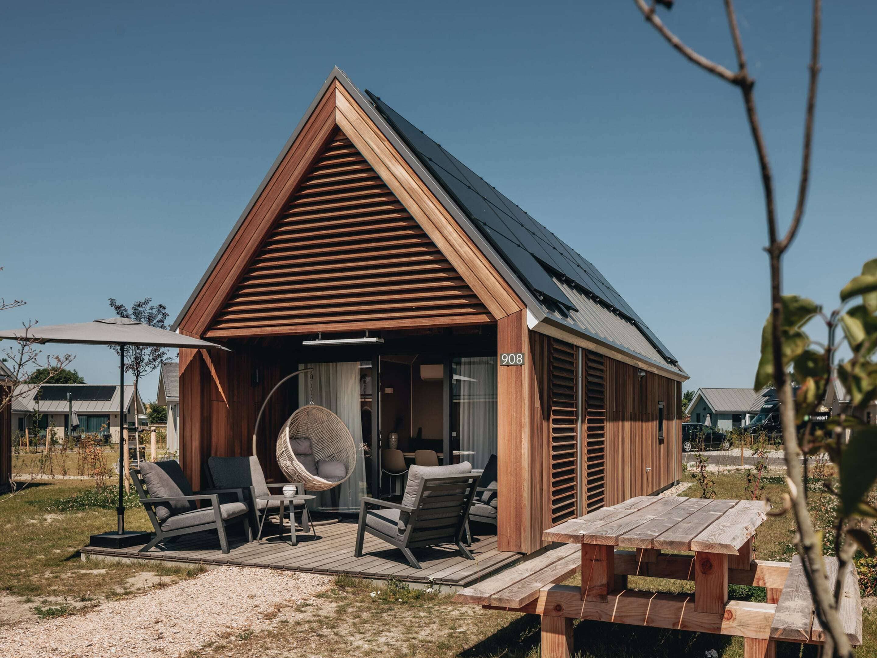 Una moderna cabina in legno con tetto spiovente, un portico coperto con posti a sedere esterni e il numero '908' ben visibile.