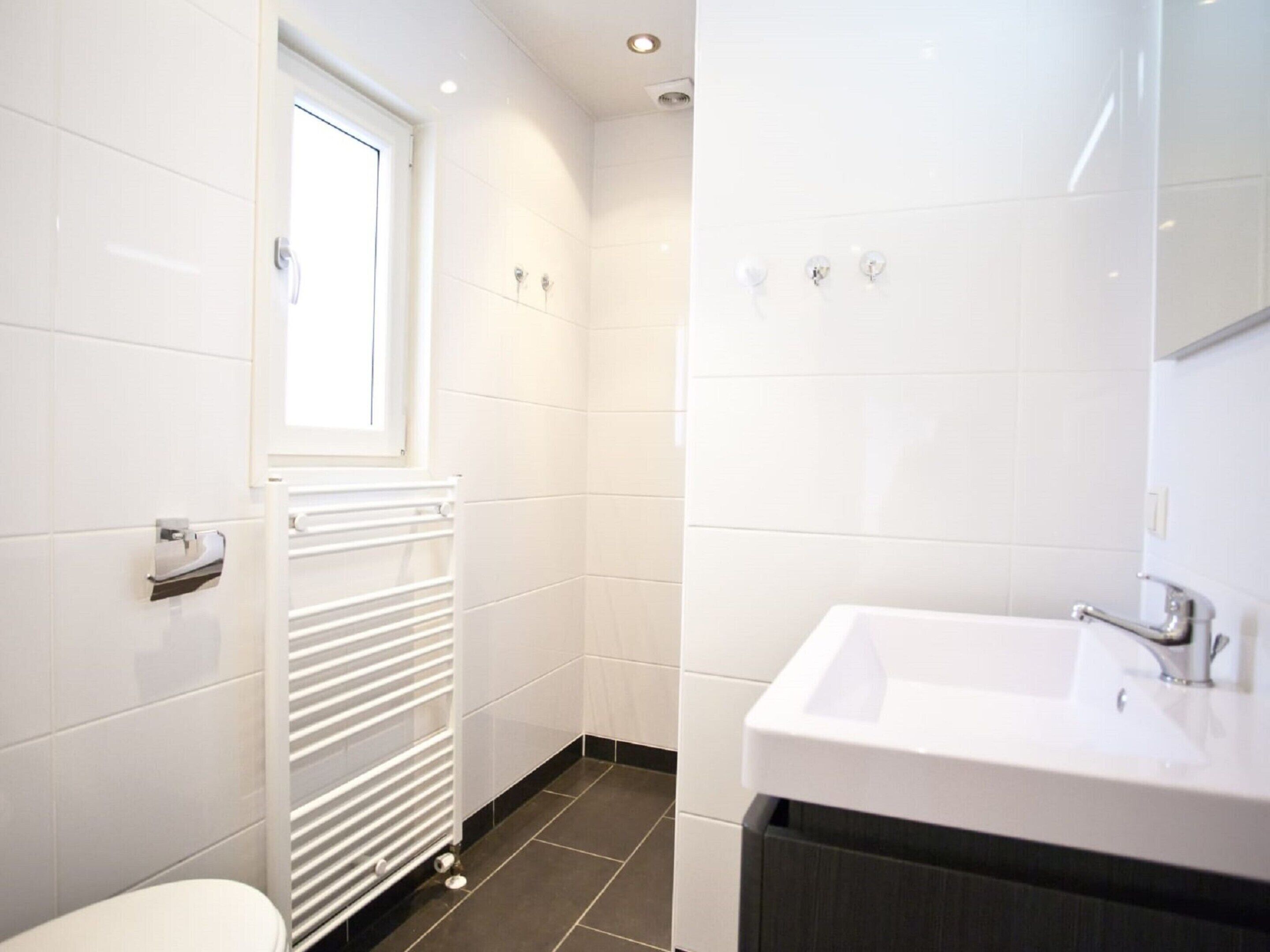 A modern bathroom with white tiles, a white radiator, a window, and a sink with a mirror.