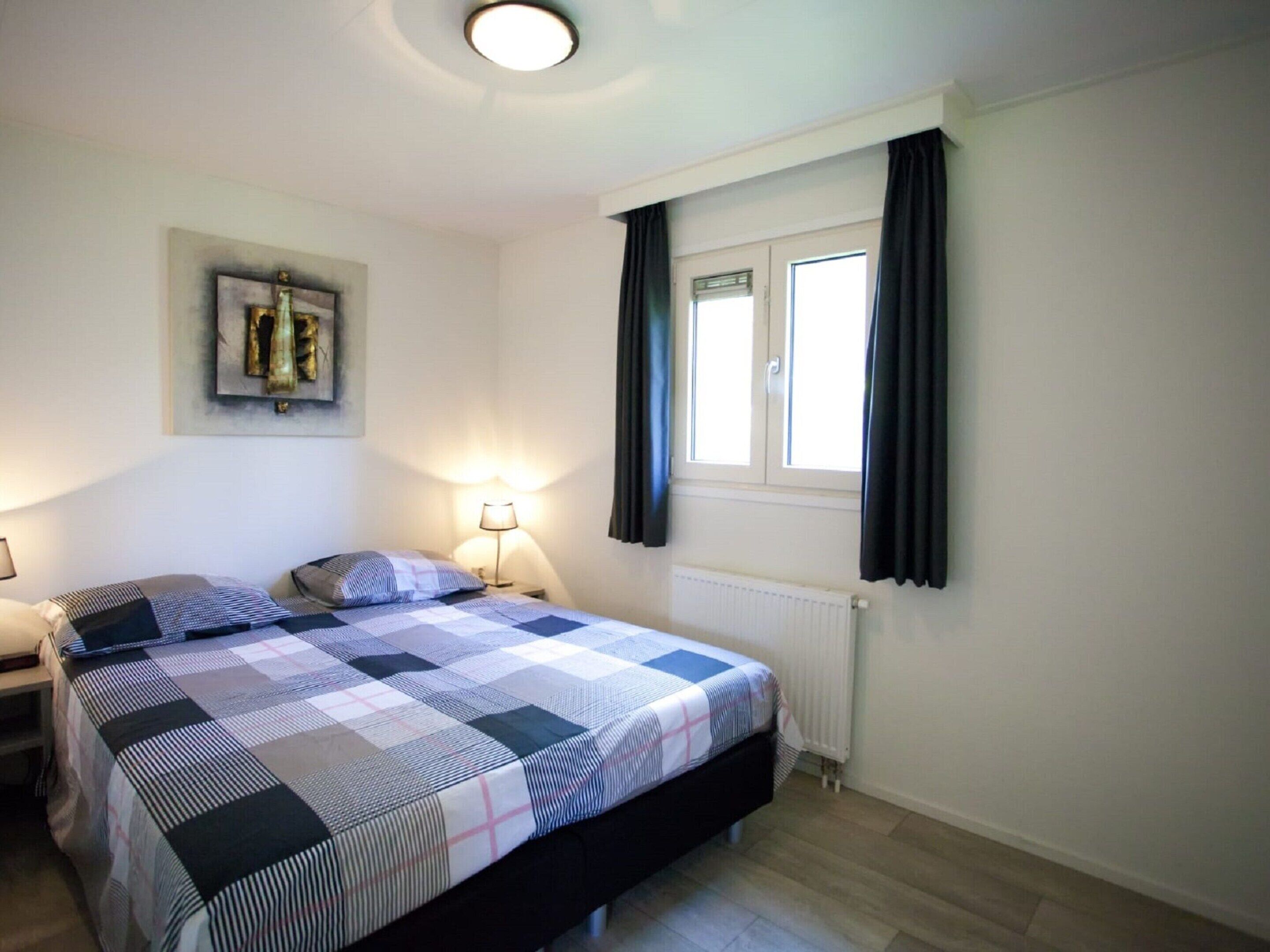 Een slaapkamer met een bed, twee nachtkastjes, een radiator en een kunstwerk aan de muur, plus een raam met gordijnen.