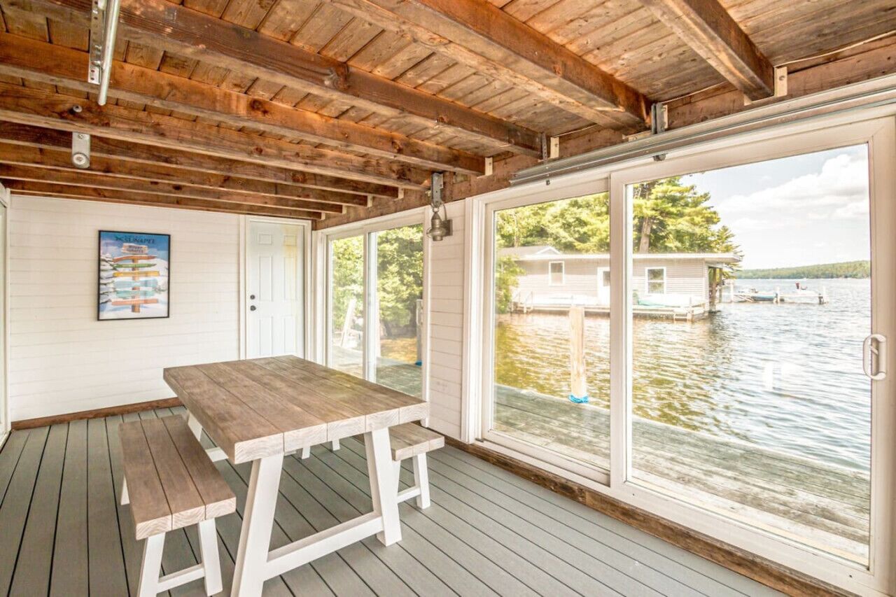A covered outdoor area with a wooden table and benches, large windows overlooking a body of water, and a poster on the wall.