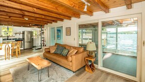 A spacious living room with a wooden ceiling, a sofa, a coffee table, and a view of a body of water through large sliding doors. - Overwater Bliss | Private Dock + 4 Min to Sunapee (Newbury)