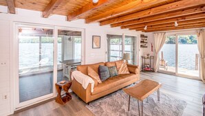 A spacious living area with a wooden ceiling, large windows, a sofa, and a small table. - Overwater Bliss | Private Dock + 4 Min to Sunapee (Newbury)