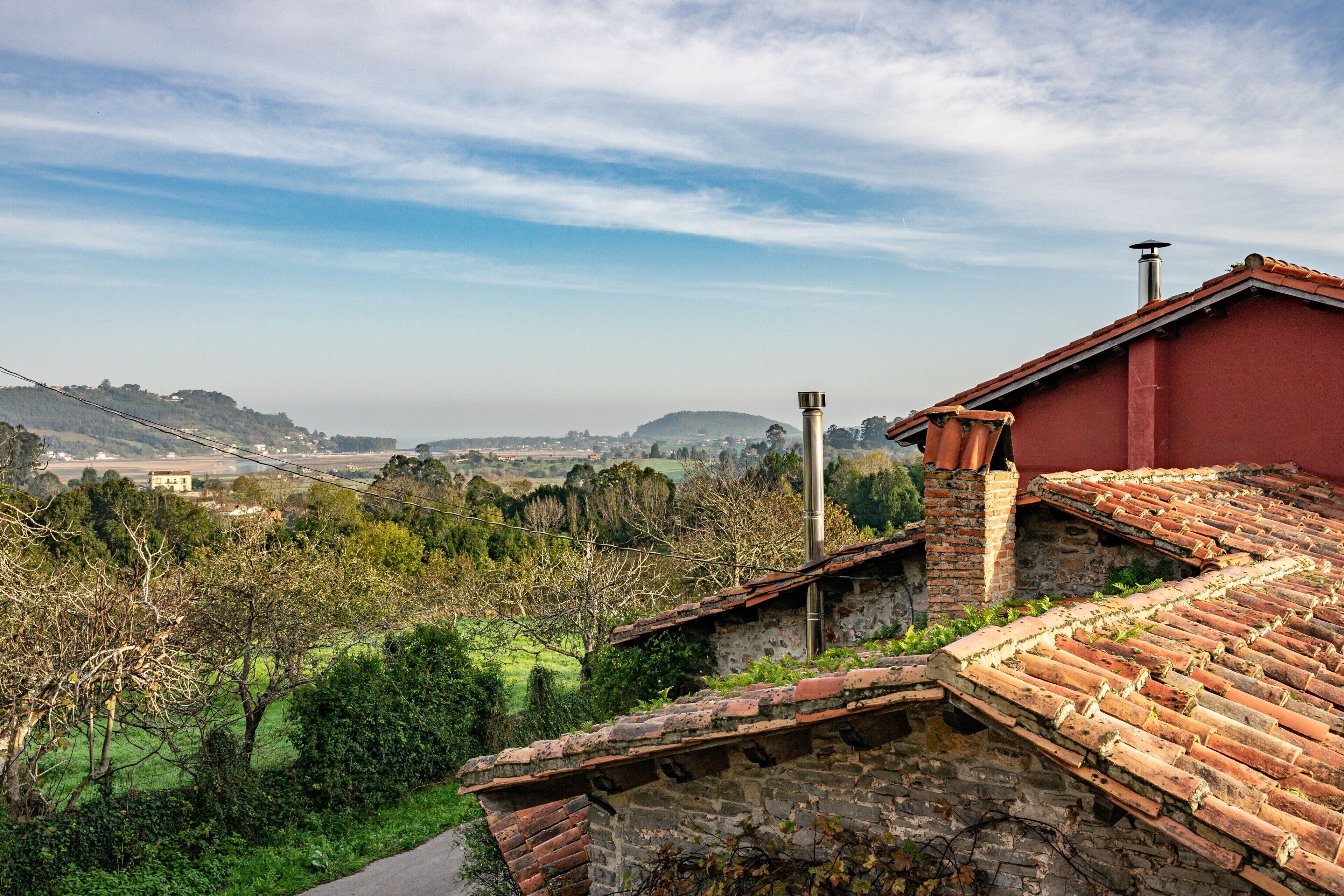 Un edificio de techo rojo con muro de piedra, una chimenea y una vista de un paisaje verde con árboles y colinas a lo lejos.